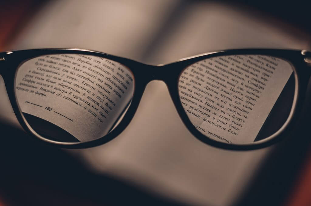 Imagem de óculos acima de um livro. O conteúdo do livro fica visível através das lentes do óculos, representando a busca por conhecimento.